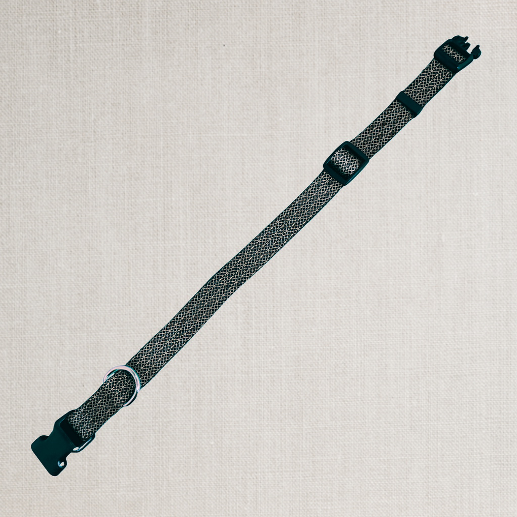 A dark gray and black patterned woven strap with black plastic buckles, laid diagonally on a light beige textured linen background.