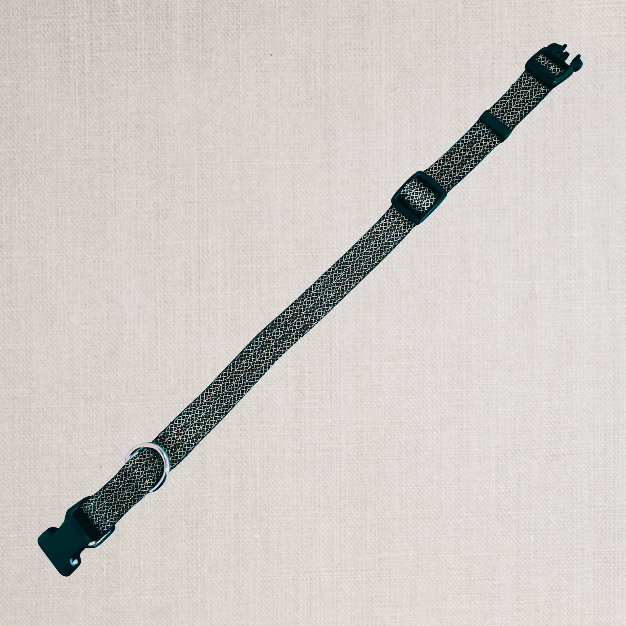 A dark gray and black patterned woven strap with black plastic buckles, laid diagonally on a light beige textured linen background.