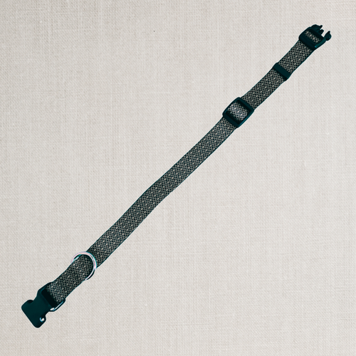 A dark gray and black patterned woven strap with black plastic buckles, laid diagonally on a light beige textured linen background.